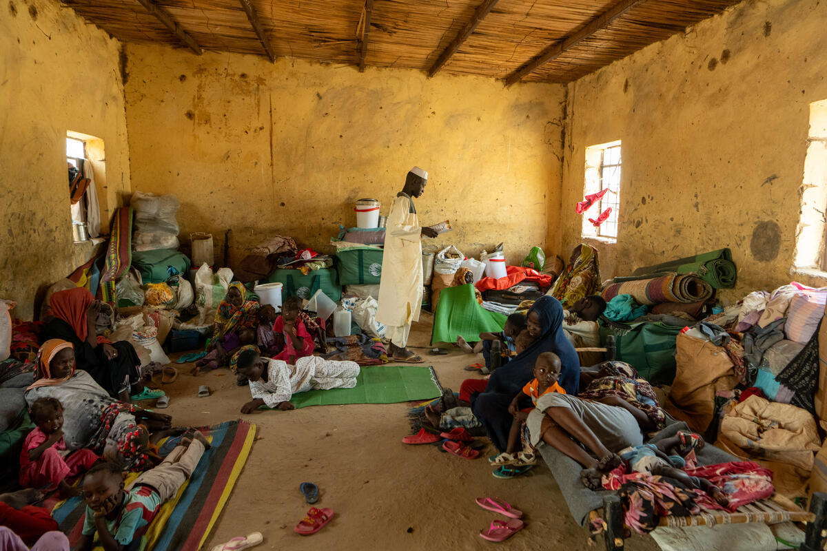 A young man stands reading while women and children sit or lie in a room crammed with their belongings.