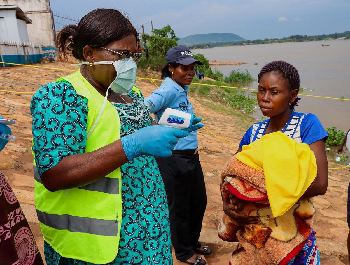 A woman wearing a mask prepares to take another woman's temperature next to a river, while a police officer stands nearby.