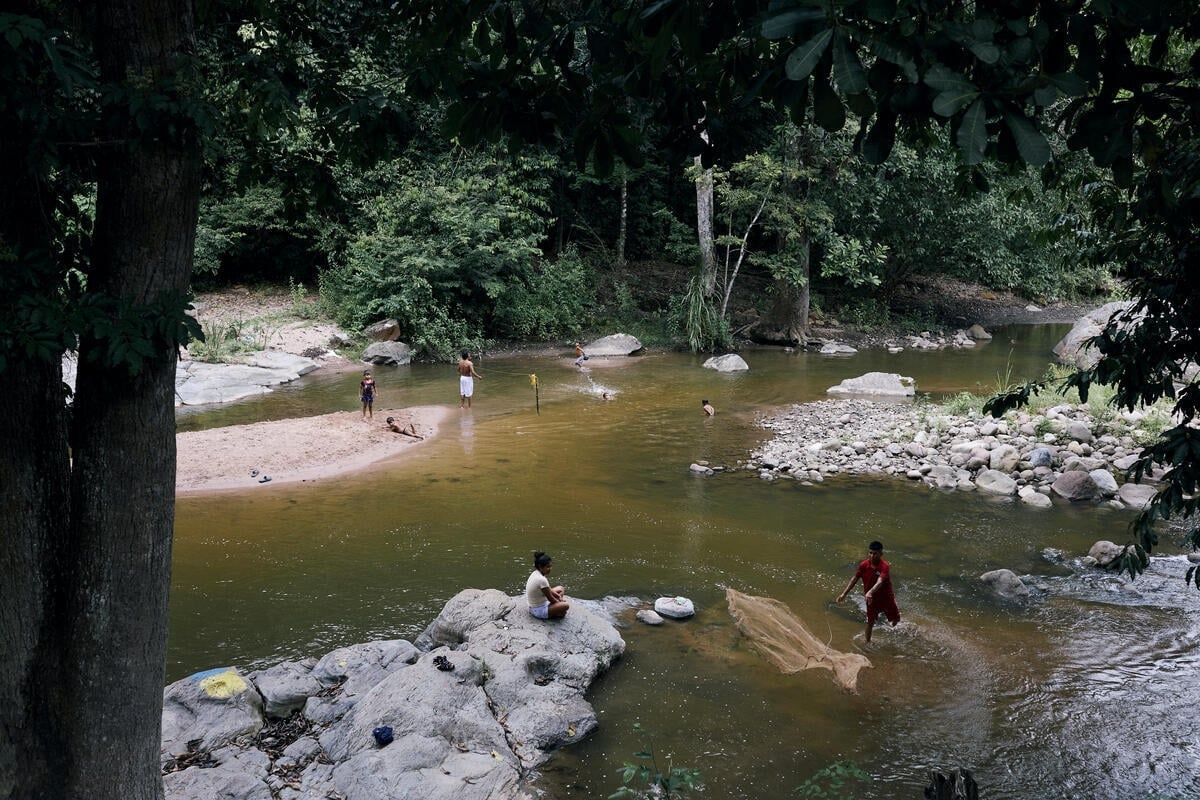 An aerial view of a river shows a man throwing a fishing net and children swimming nearby as others look on from the banks.