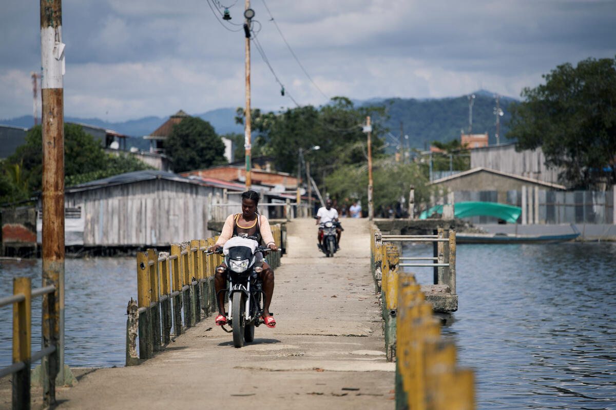 A woman woman drives a motorcycle across a walkway over a bay.