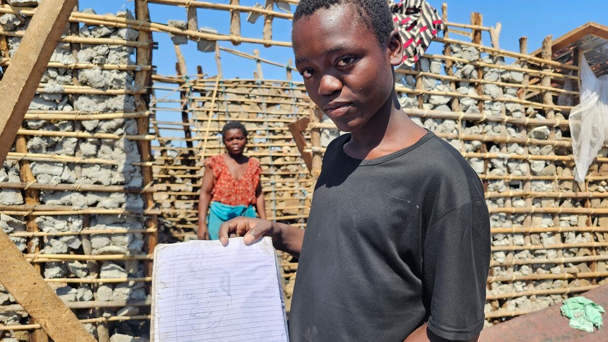 A boy holds up a note book in front of a damaged house and a woman in the background.