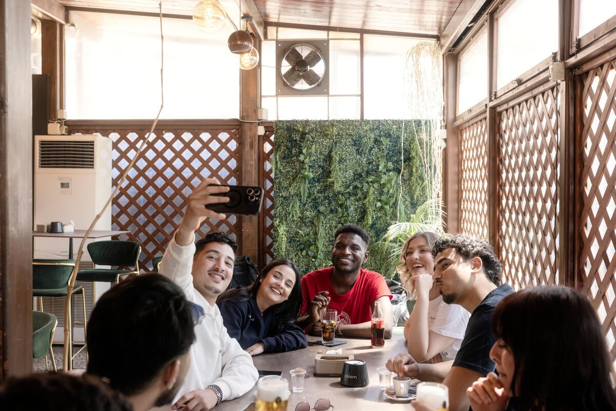 A man holds a mobile phone up for a selfie with other people seated around a table in the covered terrace of a bar 