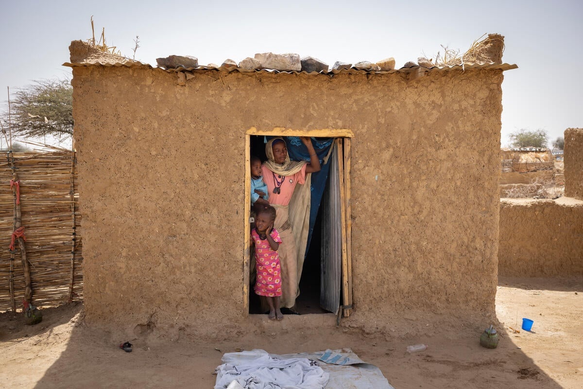 A mother stands with two children in the open doorway of a small mud brick house