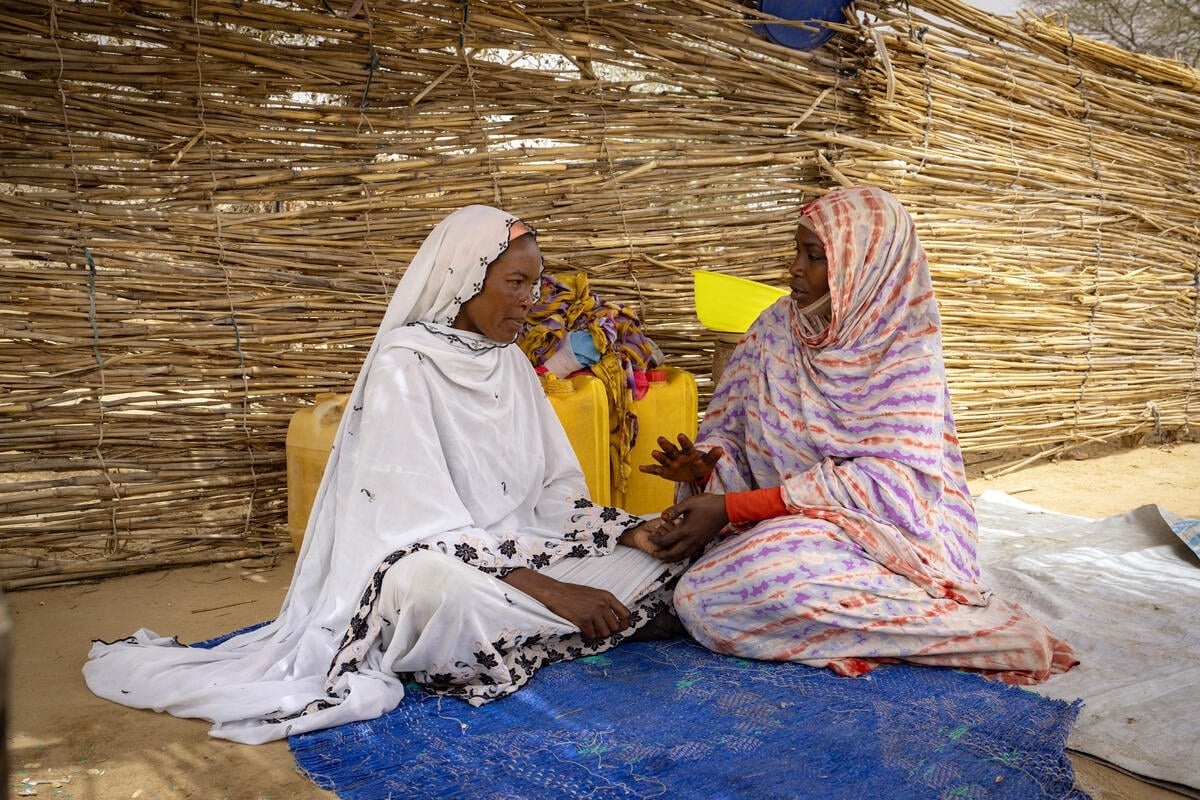 Two women sit talking in front of a woven reed wall