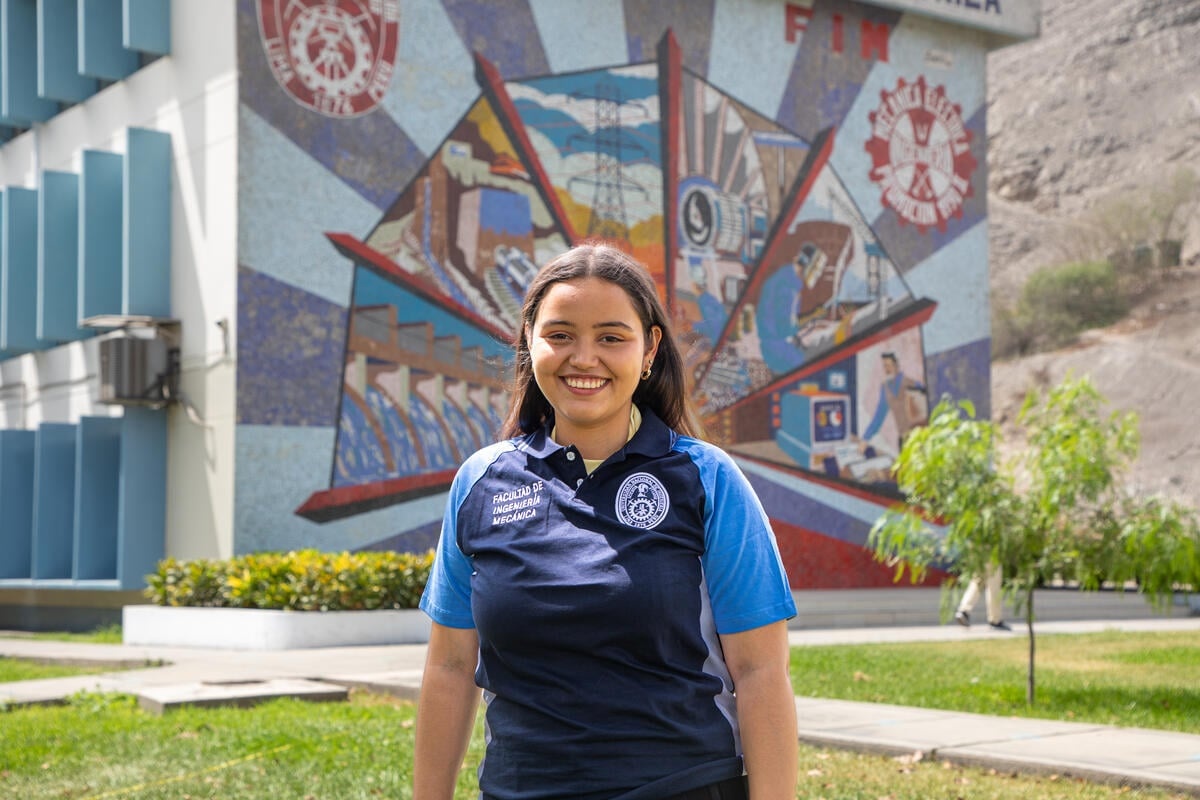A smiling young woman in a faculty polo shirt stands outside in front of a large, colourful mural painted on the side of a building