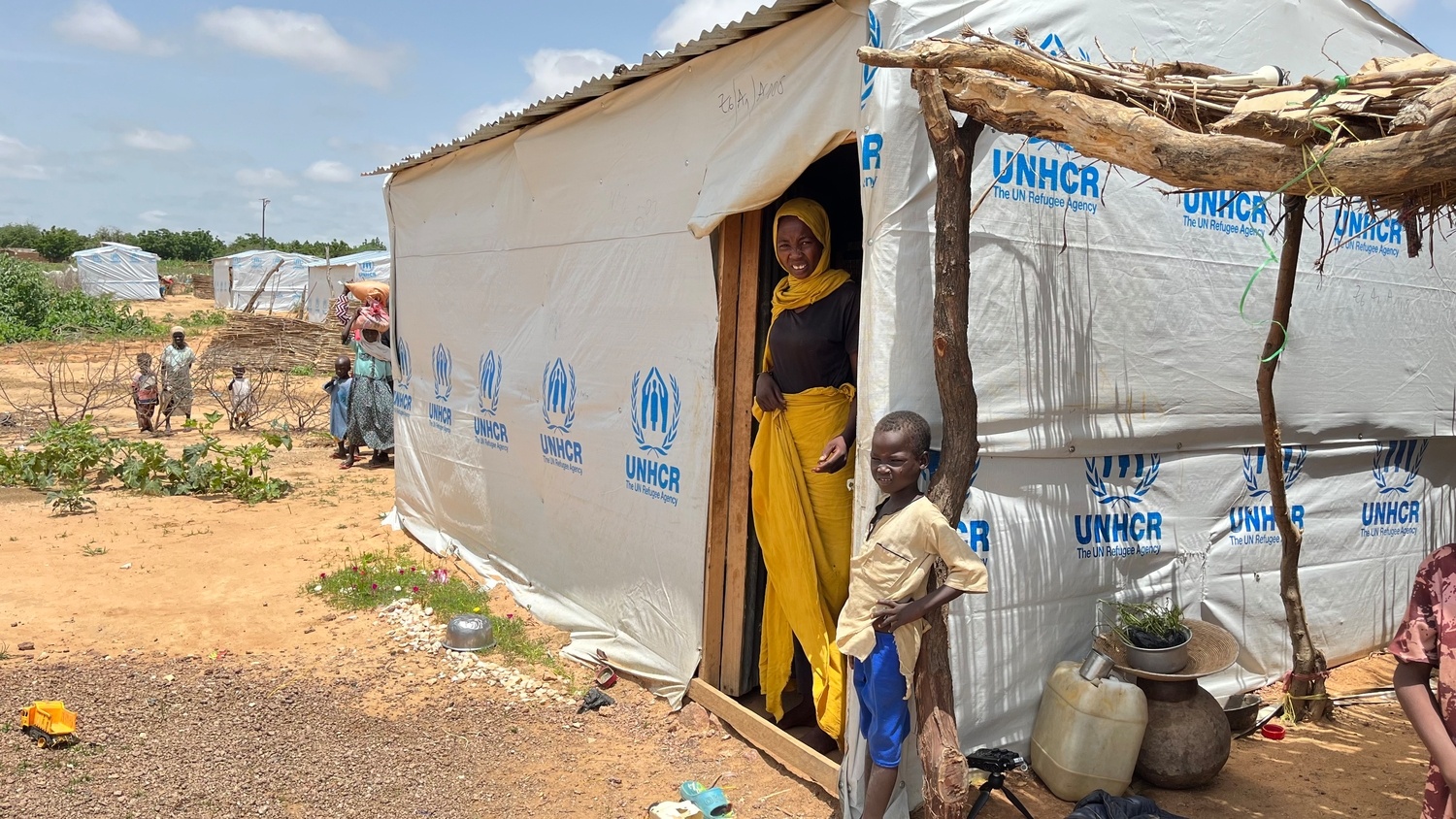 A woman and a young boy stand at the doorstep of a shelter covered in UNHCR-branded tarpaulin.
