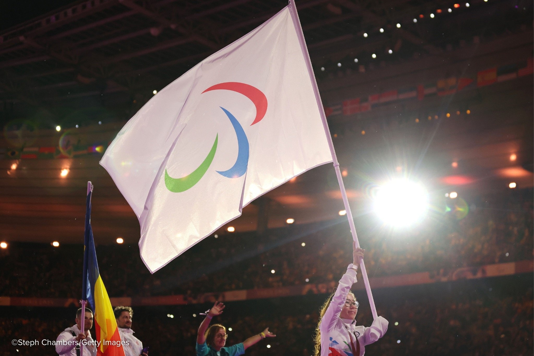 A woman wearing glasses holds a large IPC flag above her head in a brightly lit stadium.