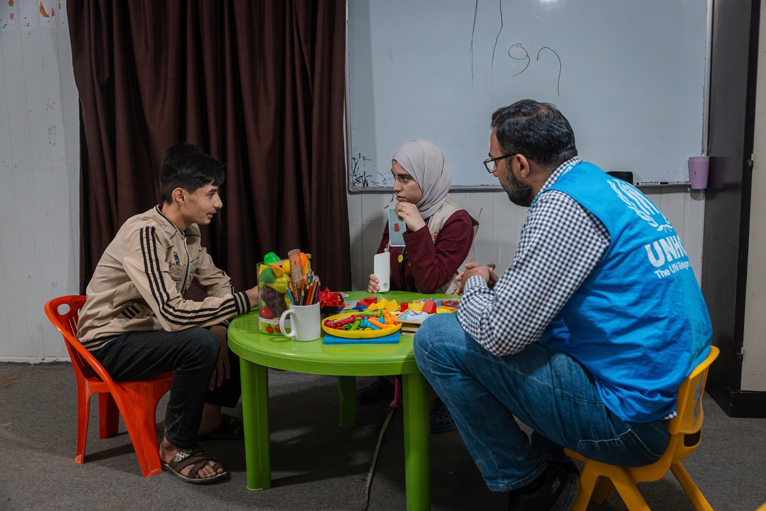 Seated on small plastic chairs at a green table, a young man speaks to a woman while a UNHCR staff member looks on.