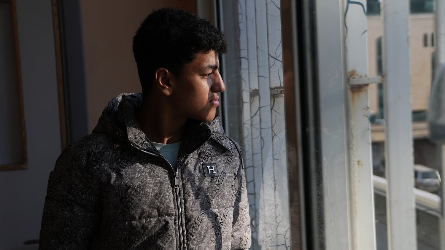 A young man stands next to a window looking out onto a courtyard