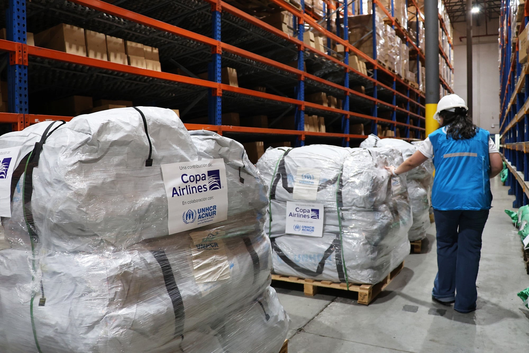 De pie junto a pallets con suministros de emergencia etiquetados con logos de ACNUR y COPA Airlines, una mujer con casco que viste chaleco azul de ACNUR