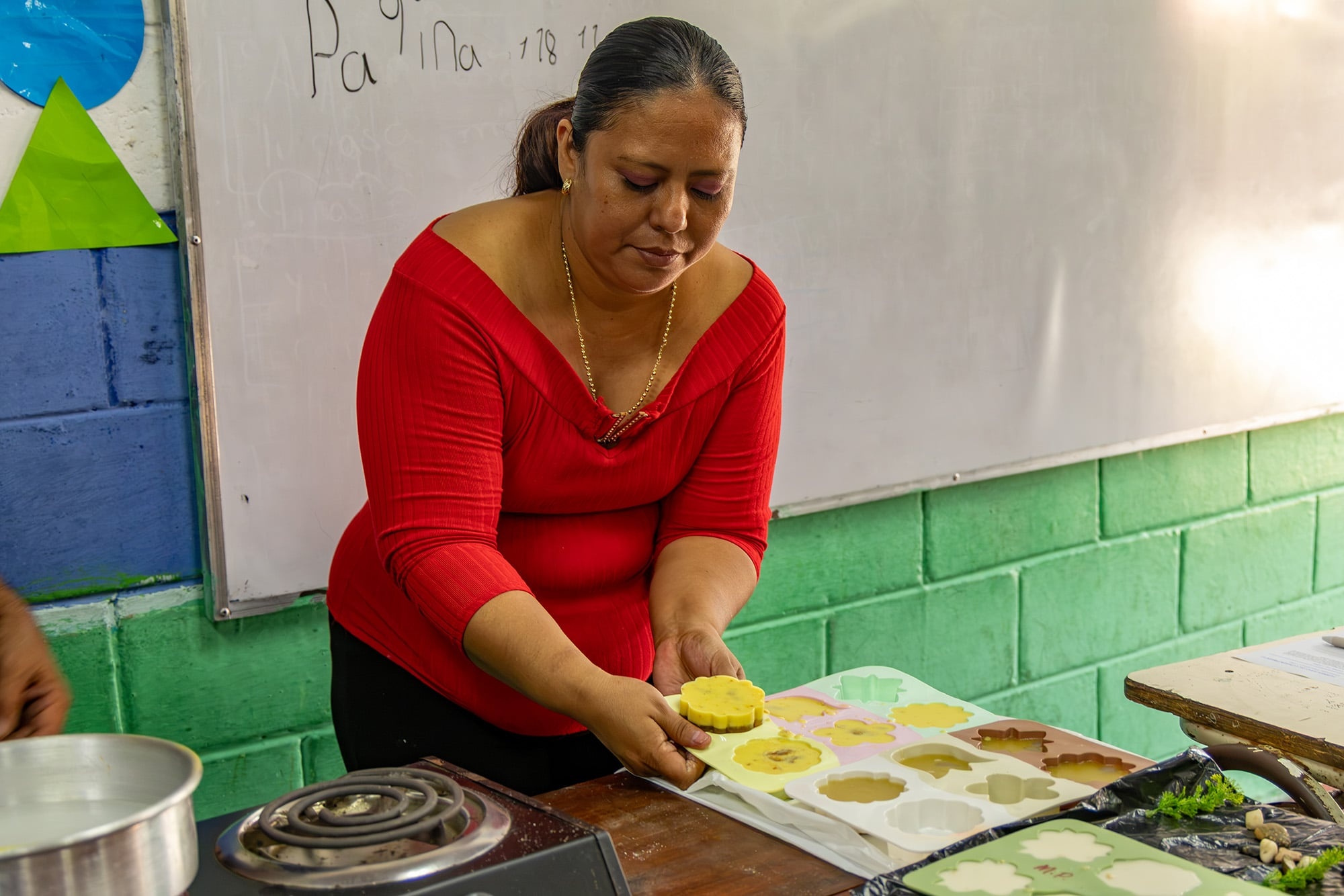Una mujer que viste blusa roja, desmolda con sus manos un jabón amarillo dentro de un aula