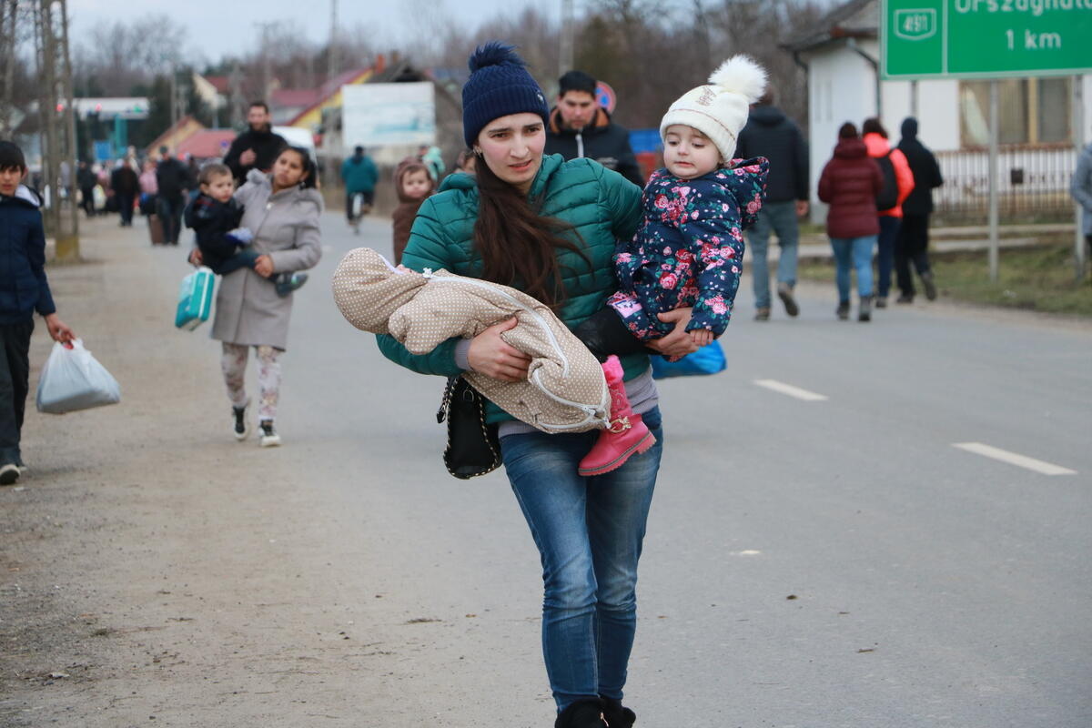 A young woman walks along the side of a road carrying a toddler and a baby in her arms 