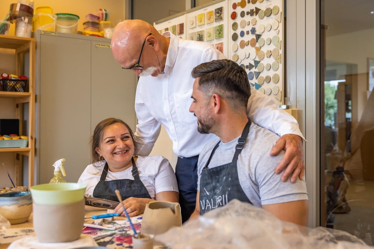 A man embraces a couple wearing aprons in a ceramics workshop.