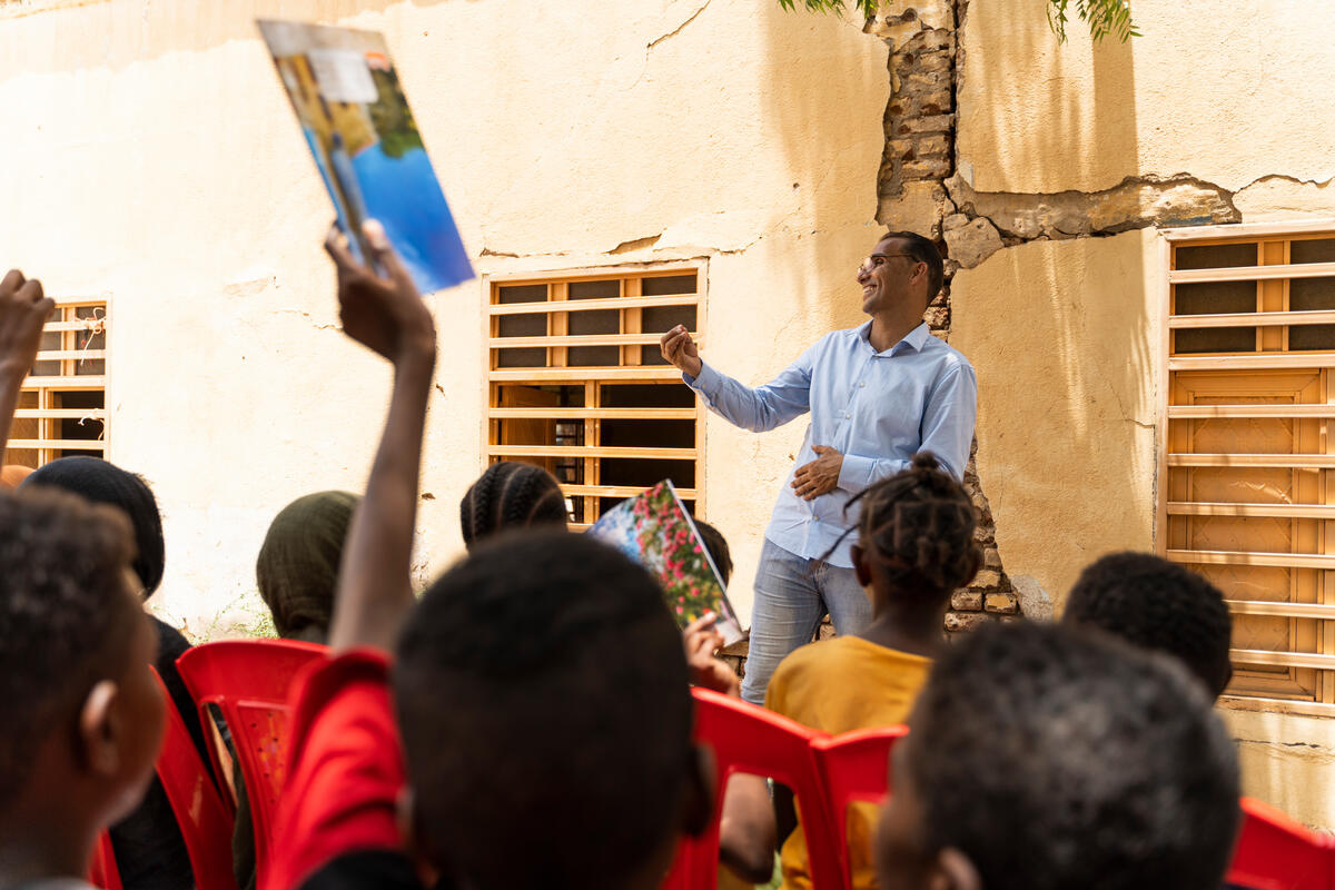 A smiling teacher stands outside in front of a group of students seated in red chairs.