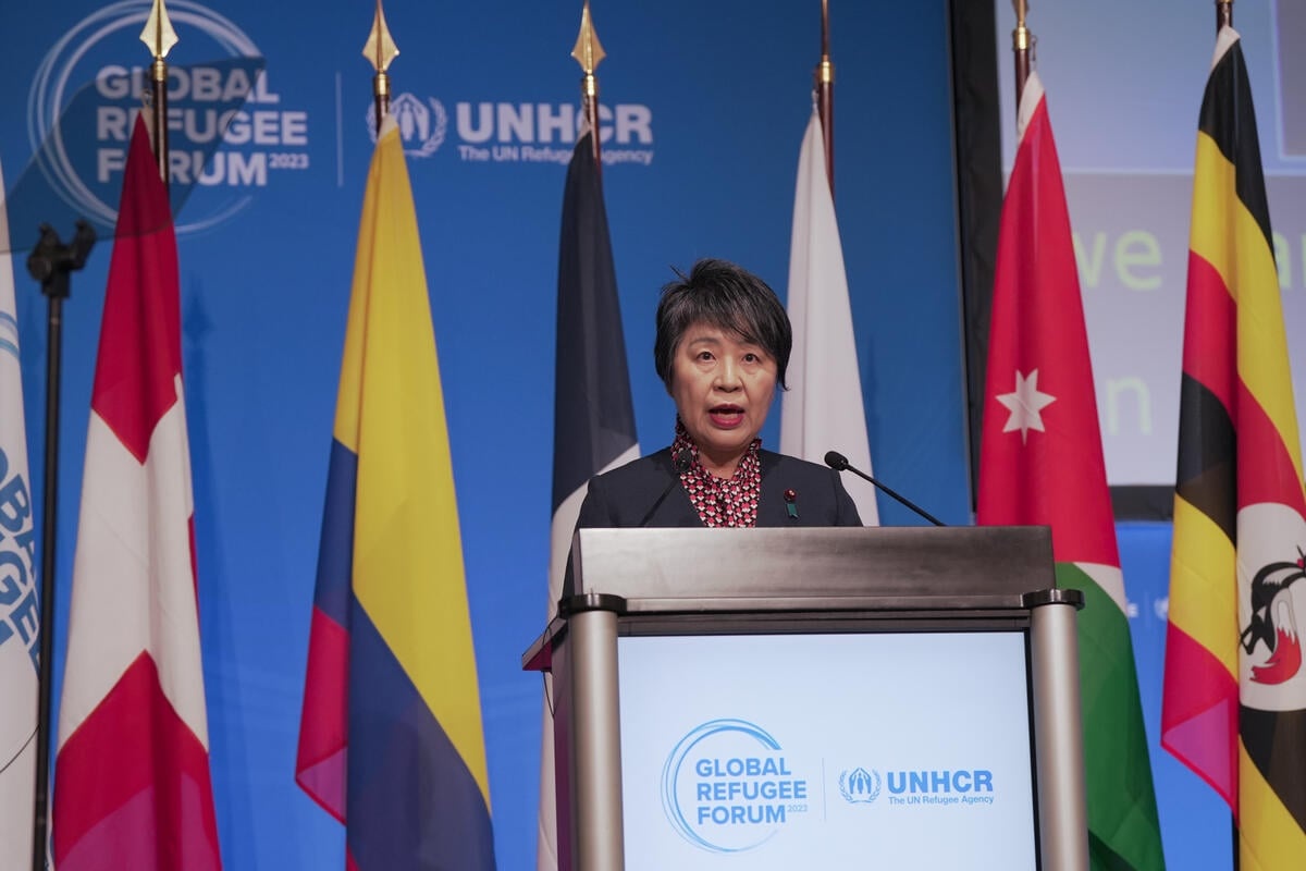 Kamikawa Yoko speaks at a podium with many flags behind her.