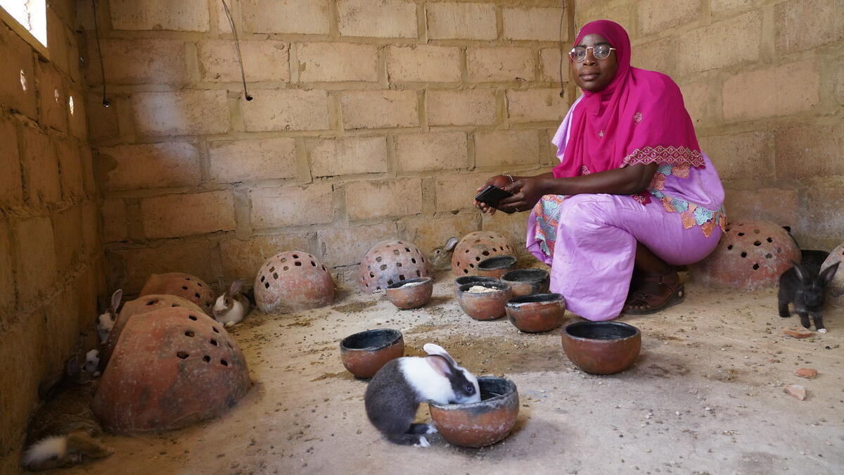 A woman in a pink headscarft sits in a brick walled outbuilding with rabbits feeding from bowls
