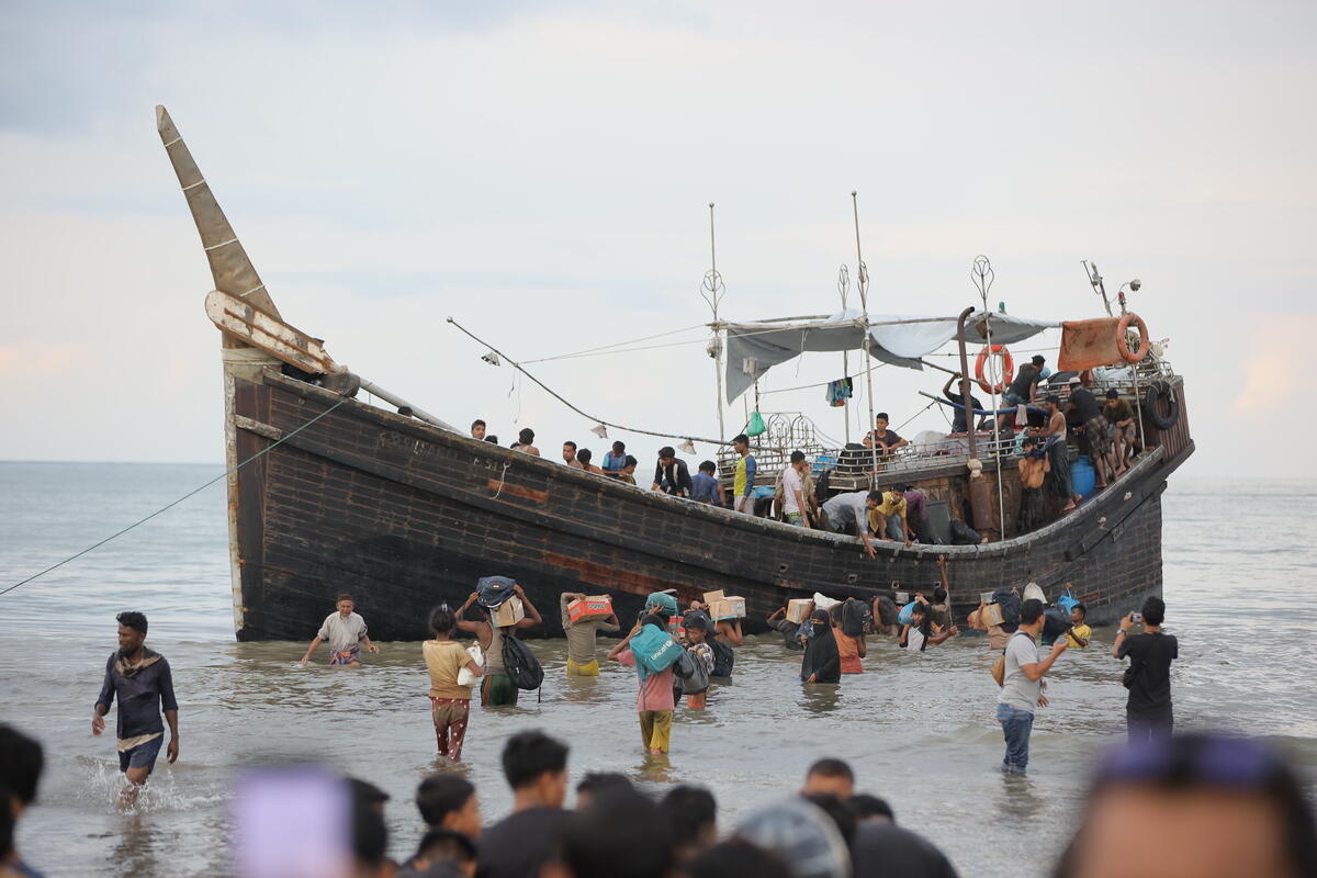 A group of people carrying belongings stand in the shallows alongside a wooden boat carrying refugees.