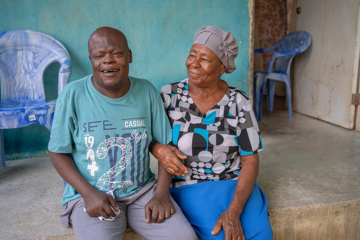 An older woman in a headscarf sits on a porch linking arms with a younger man.