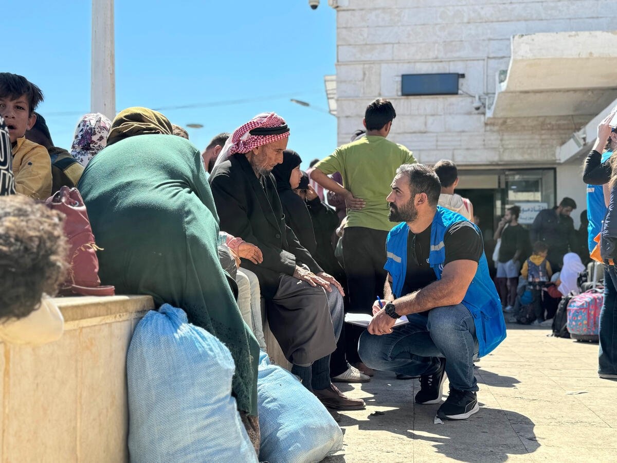 A UNHCR staff member with a notebook crouches down to talk to a seated elderly man. 