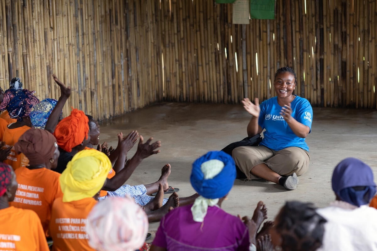 A woman in a UNHCR branded t-shirt sits on the floor leading a group of women in clapping.
