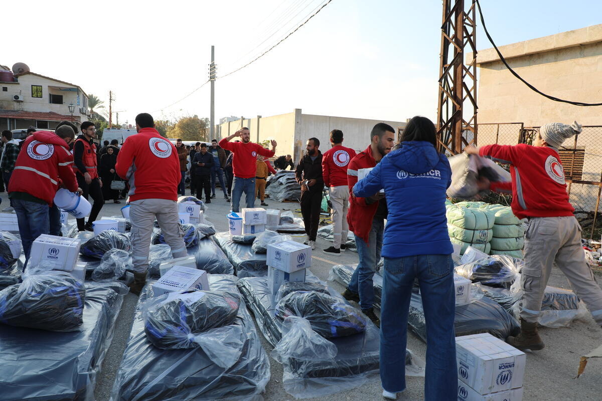 A woman in a blue UNHCR jacket hands a box to a man in a red jacket during a distribution of emergency aid items.