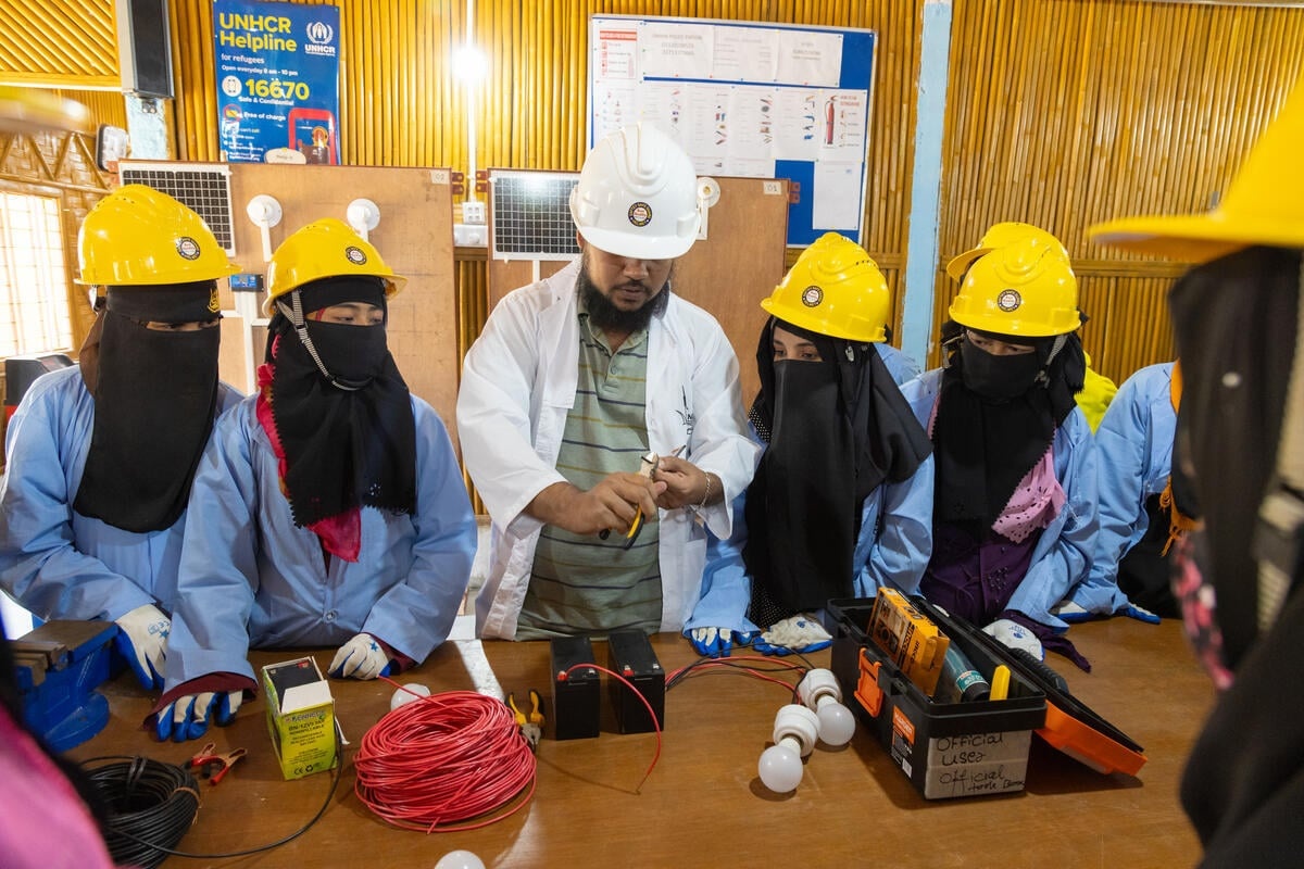 Women wearing niqabs and hard-hats watch as a man in a white coat and hard-hat demonstrates cutting a wire.