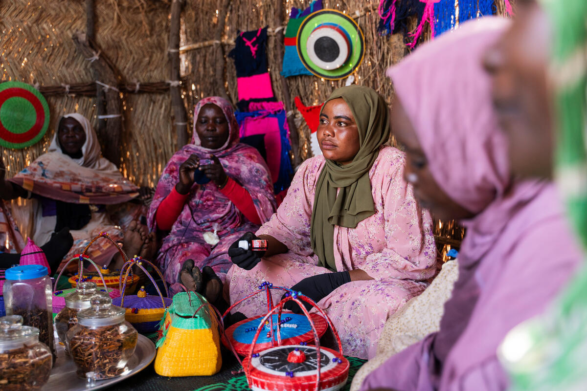 Women sit on the floor of a shelter showing off crafts they have made.