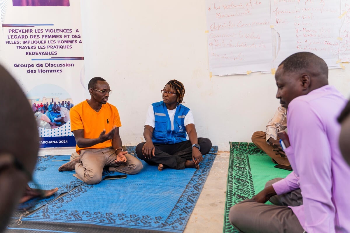 A woman in a UNHCR vest sits cross-legged on a mat listening to a man in an orange t-shirt talking while others look on.