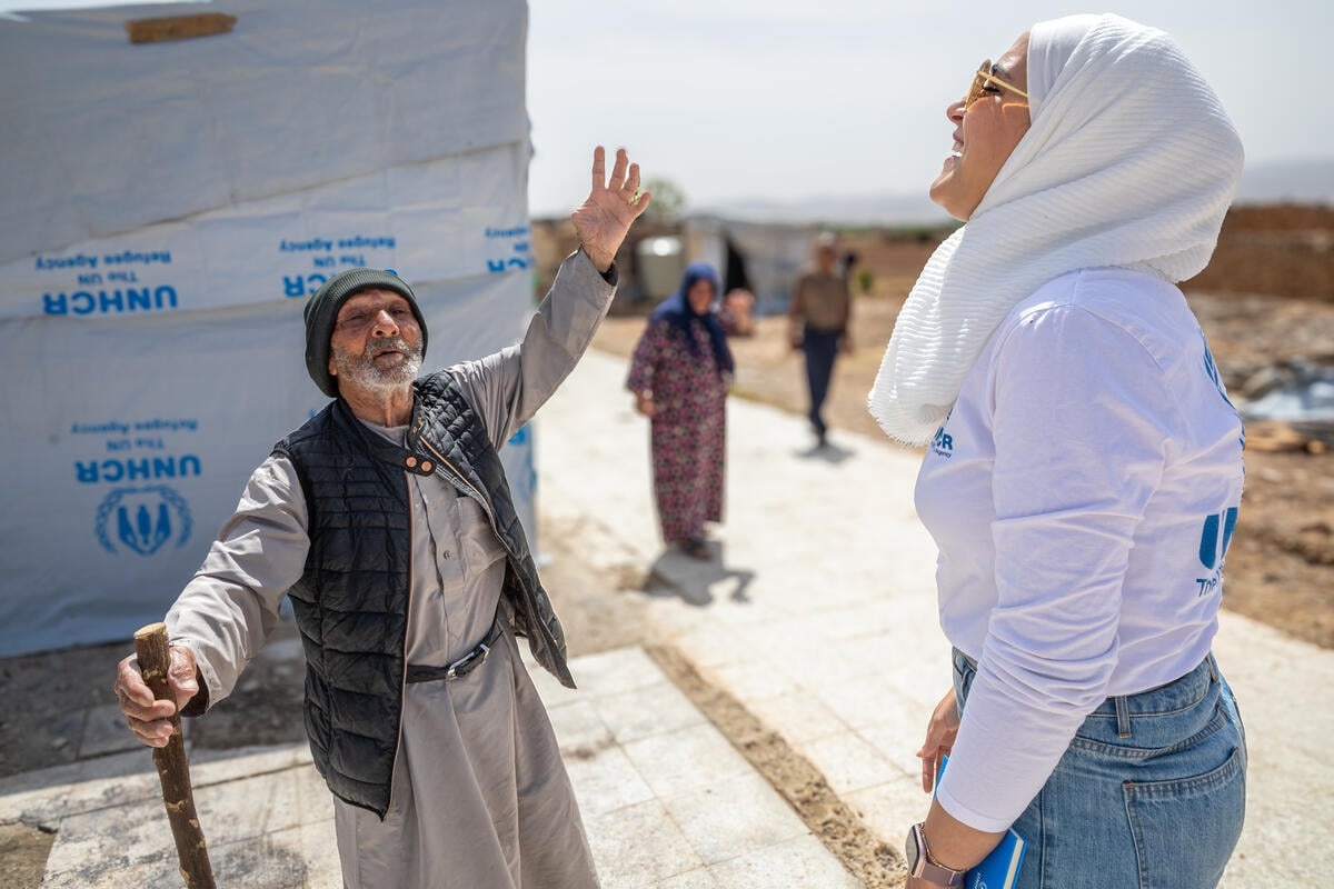 An elderly man holding a walking stick talks animatedly to a member of UNHCR staff outside a plastic covered shelter 