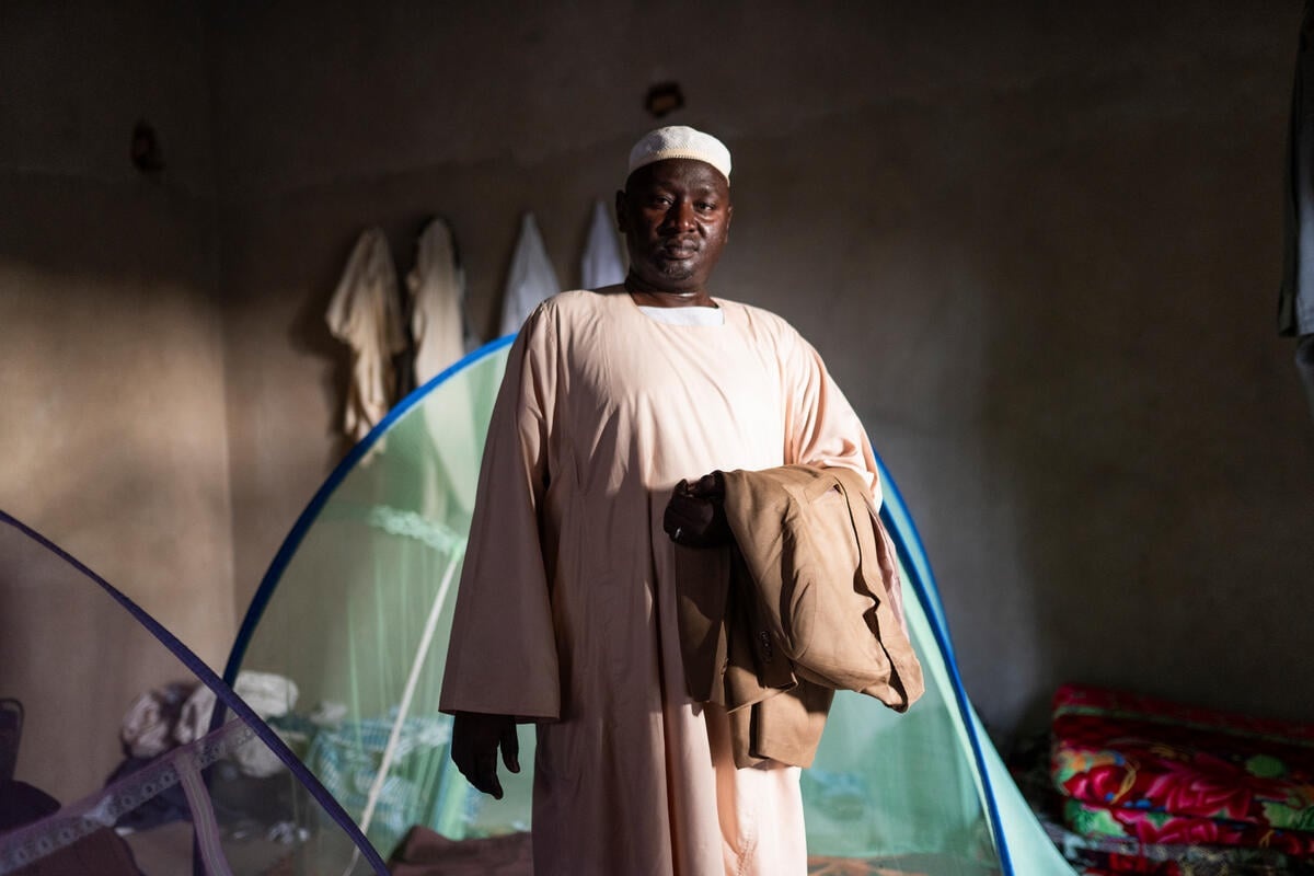 A man stands holding a folded suit over his left forearm in a shaded room with mosquito net tents in the background