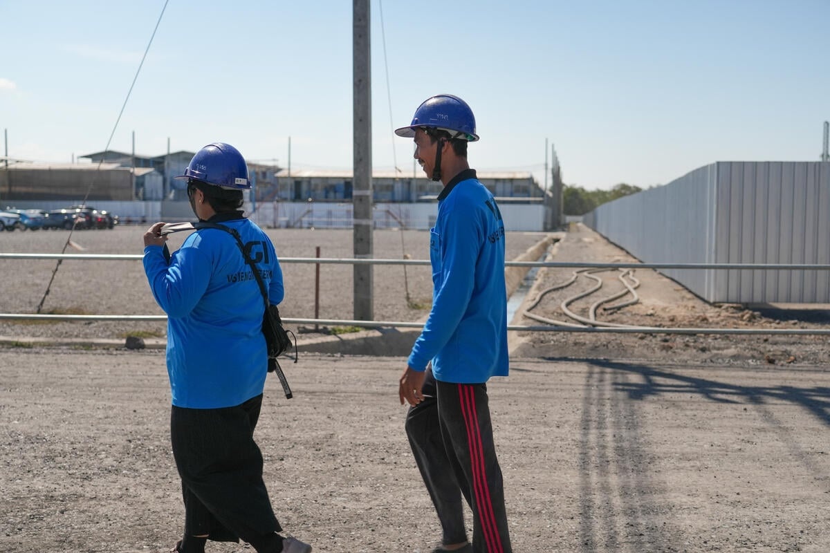A woman and a man wearing blue uniforms and hard hats walk in file down a road next to a car park