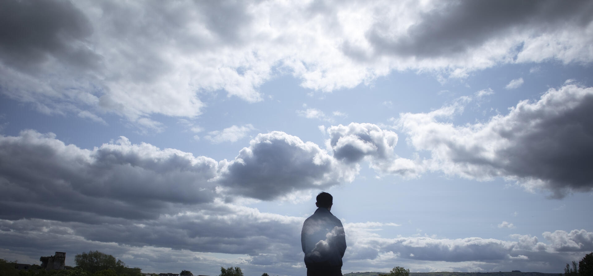 A man stands looking at the clouds in the sky. His torso fades into the scenery. 