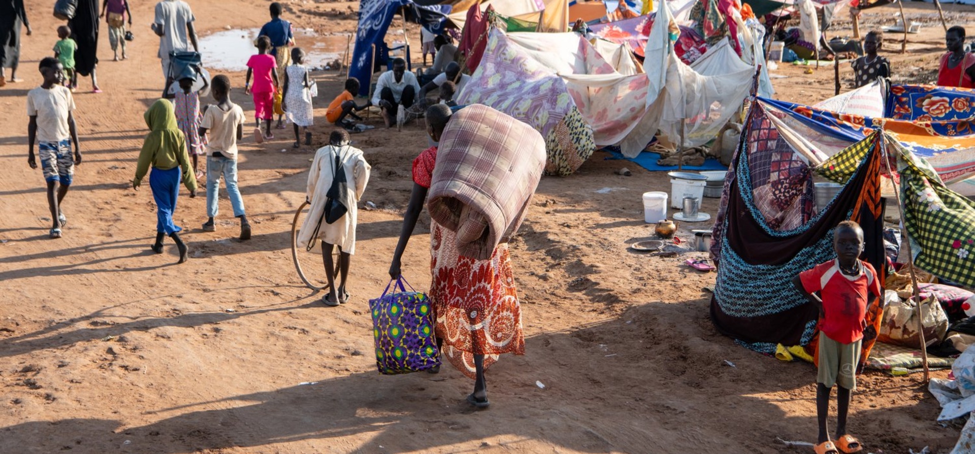 A woman carrying a blanket and a bag and several children arrive at a makeshift settlement.