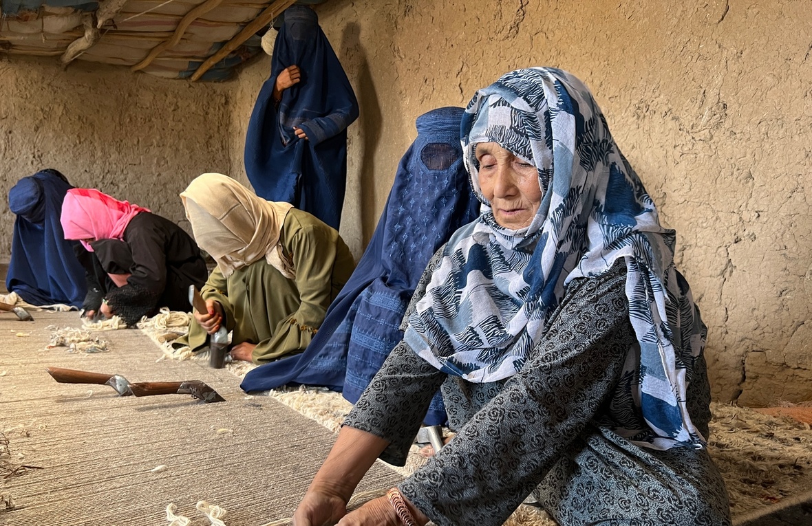 Several women sit on the floor of a hut weaving a large carpet.