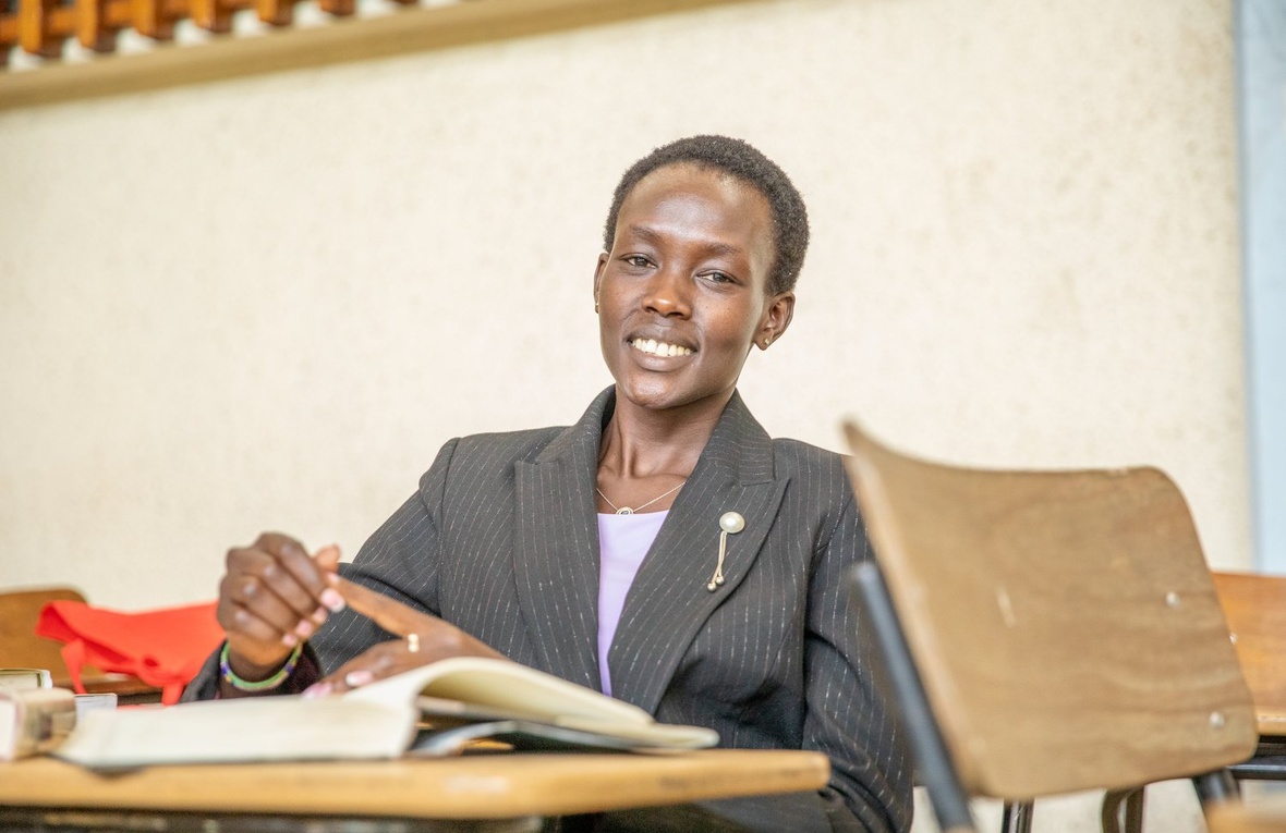 A woman in a suit jacket sits behind a desk in a lecture room.