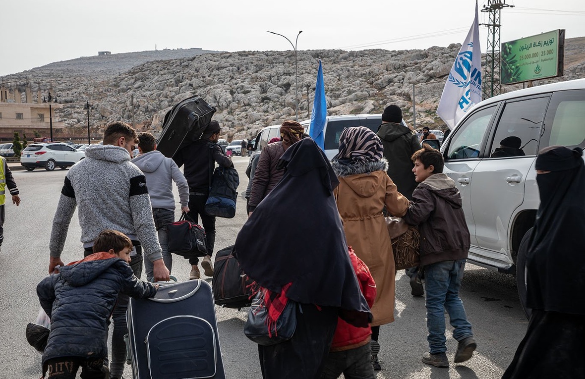 A group of people walk on a road with their belongings, towards a large rocky outcrop