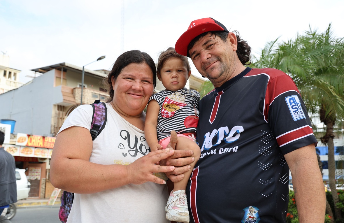 A man and a woman smile at the camera while holding their small child, buildings and a tree in the background.