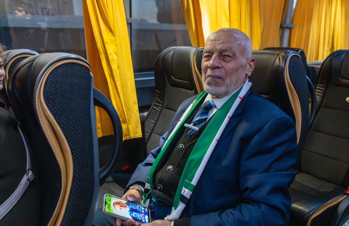 A man sits on a bus wearing a suit and scarf and holding a mobile phone