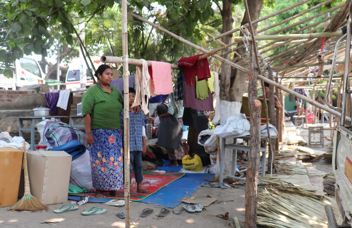 A woman stands under a tree with her arm around a young boy next to an unfinished bamboo structure and surrounded by belongings