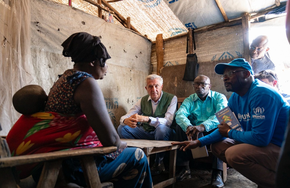 A woman with a young child strapped to her back sits opposite three men inside a tented structure