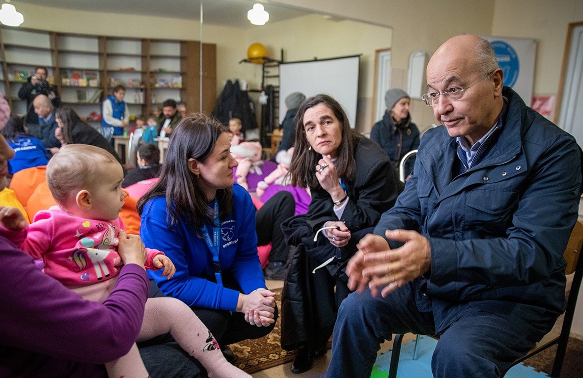A man talks to a woman with a baby girl on her lap, seated in a communal room with other people in the background