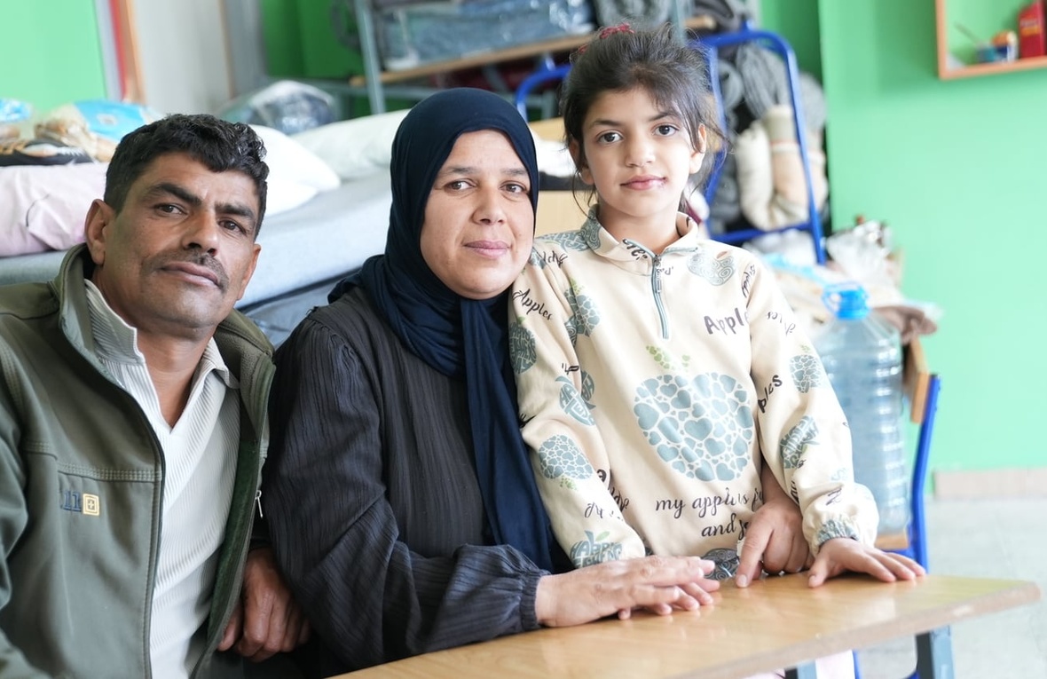 A couple and their daughter sit at a school desk in a classroom with a bed in the background 