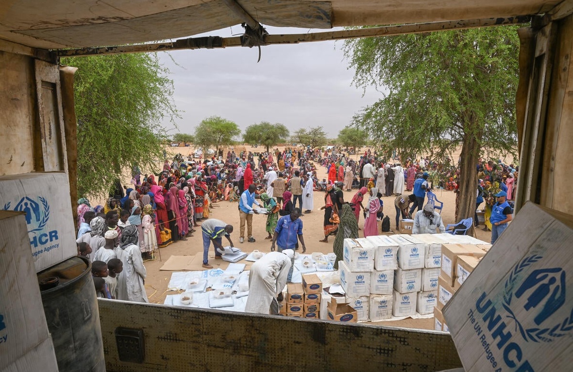 A view from the back of a truck showing workers unloading relief supplies for refugees waiting nearby.