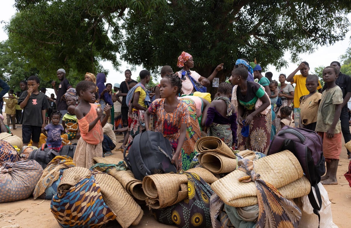 A large group of newly displaced people gather with their belongings in front of a large tree