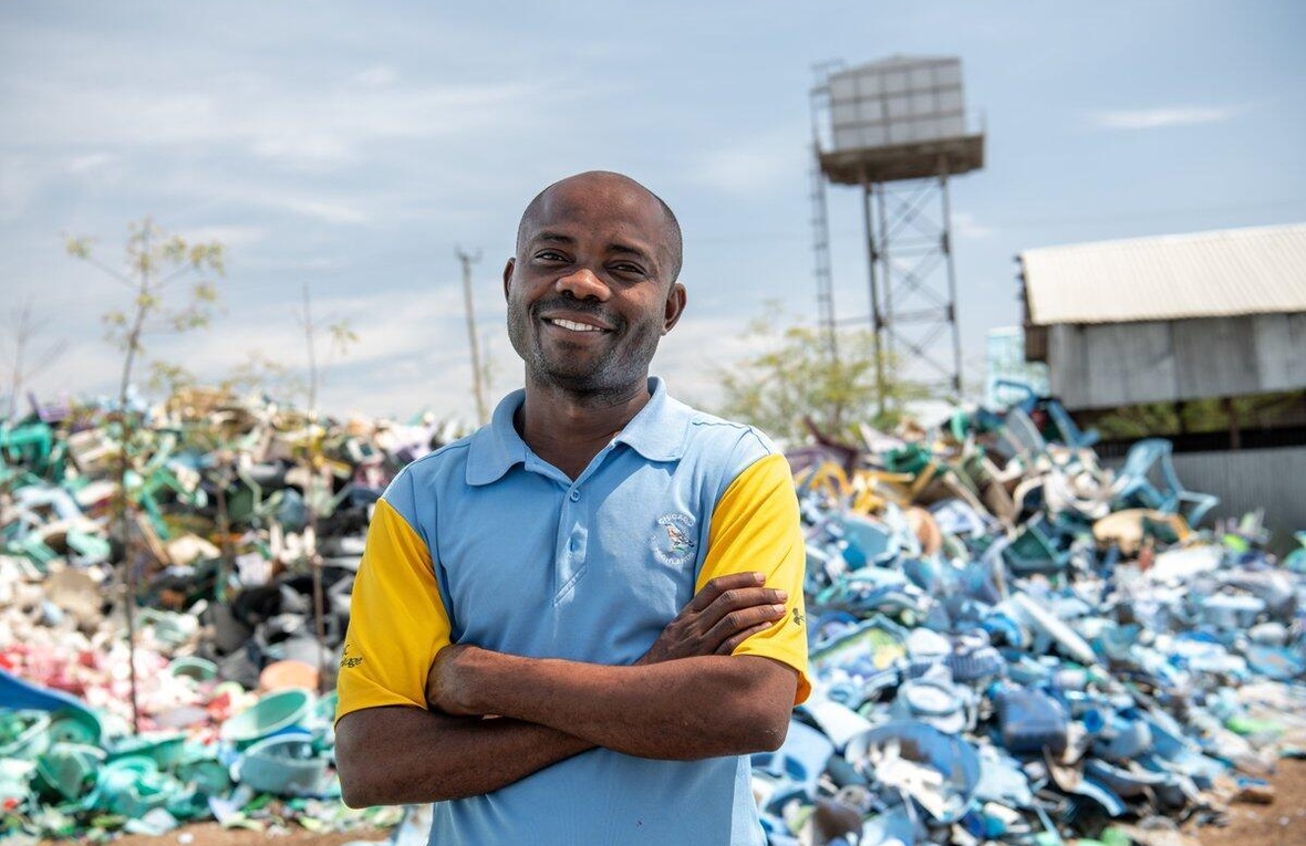 A smiling man stands in front of a huge pile of plastic waste.