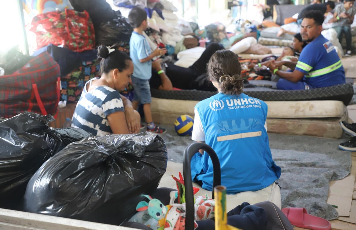 A UNHCR worker squats on the floor alongside refugees and their belongings in a temporary shelter