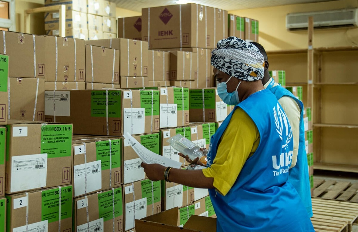 A masked UNHCR worker stands in a warehouse reviewing boxed supplies of medication