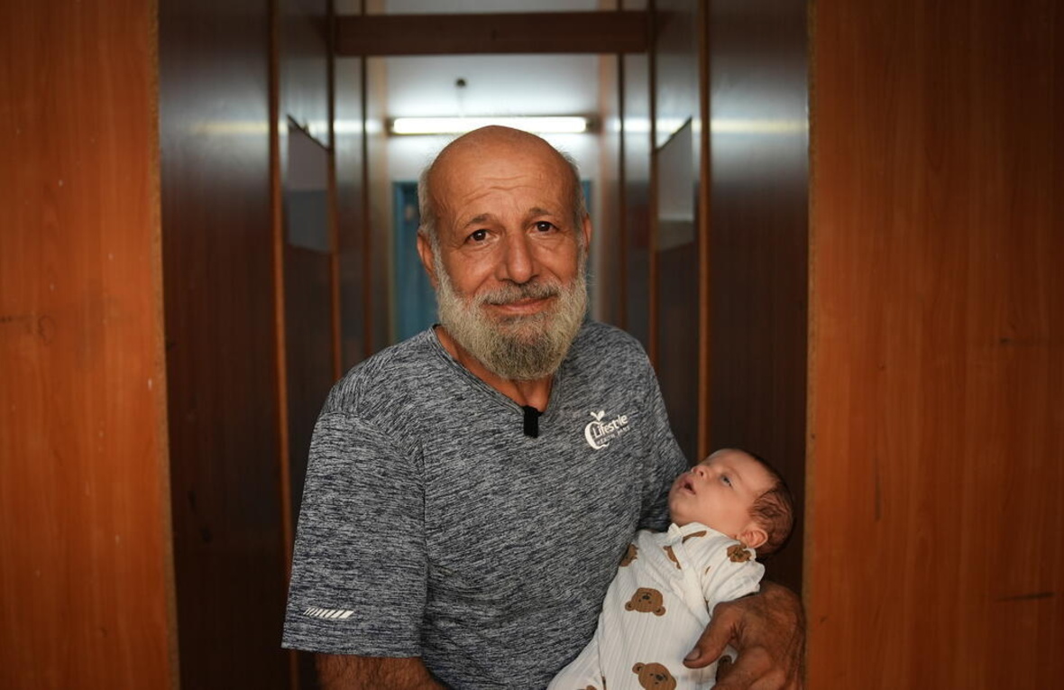 An older man with a beard holds a baby and looks into the camera.