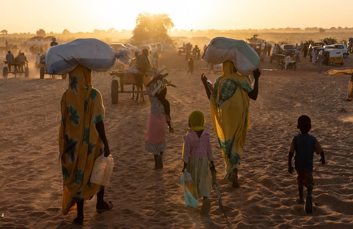 Sudanese refugees and their children walk towards the sun, carrying their belongings on their heads or on carts.