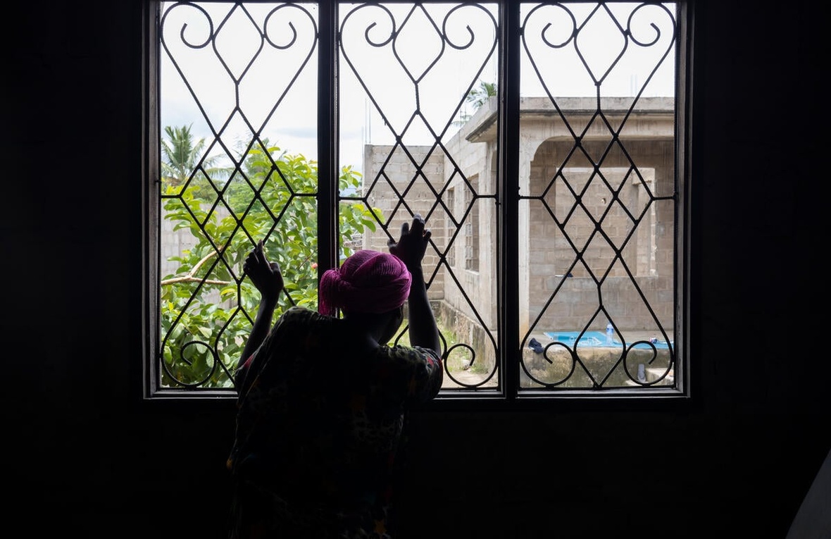 A woman with her back to the camera looks out of a window with her hands on the security bars.